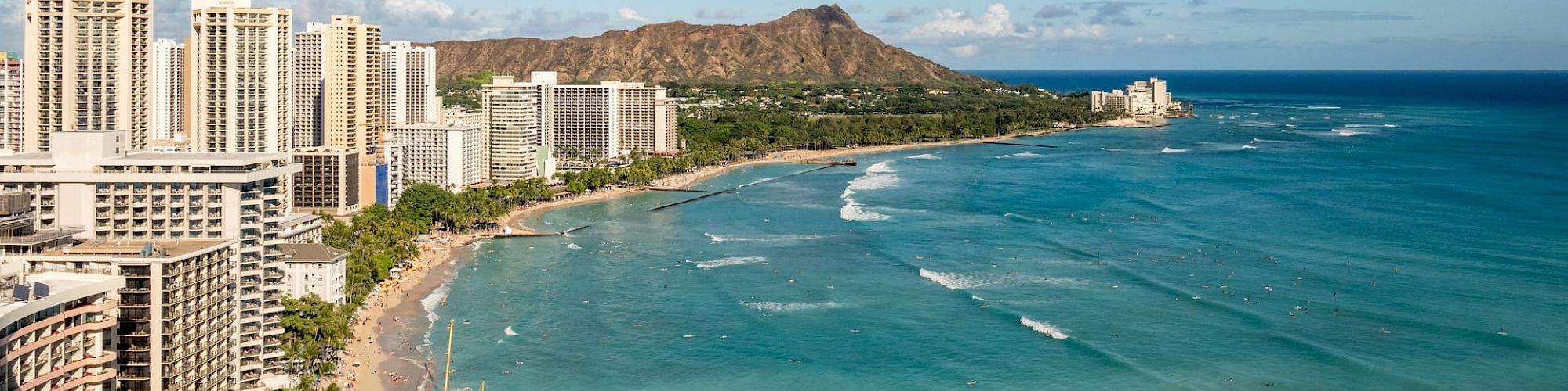 The image shows a coastal cityscape with tall buildings, a sandy beach, and a scenic mountain in the background by a turquoise ocean.
