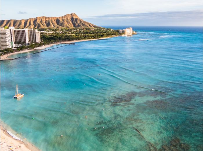 A scenic view of a coastline with clear turquoise waters, a sandy beach, buildings, and a mountainous backdrop, likely capturing a tropical location.