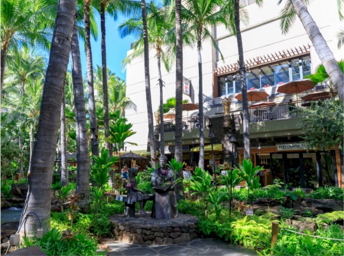 This image shows a lush, tropical outdoor area with tall palm trees, statues, and a building with balconies in the background.