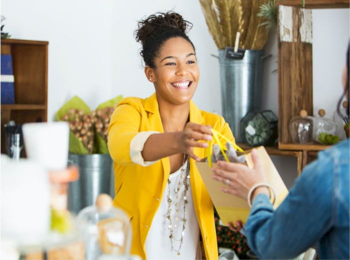 A smiling woman in a yellow blazer hands a gift bag to another person in a store with shelves and decorative items in the background.