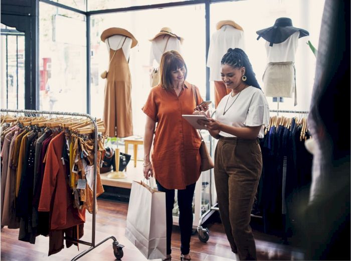Two women in a clothing store, one holding a shopping bag and the other showing a tablet, surrounded by clothes and mannequins.