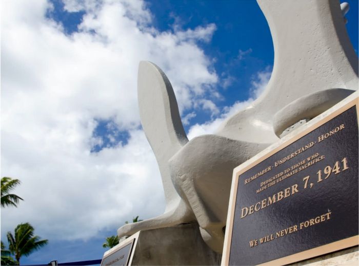 The image shows a memorial plaque dated December 7, 1941, with the inscription "WE WILL NEVER FORGET," against a background of a partly cloudy sky.