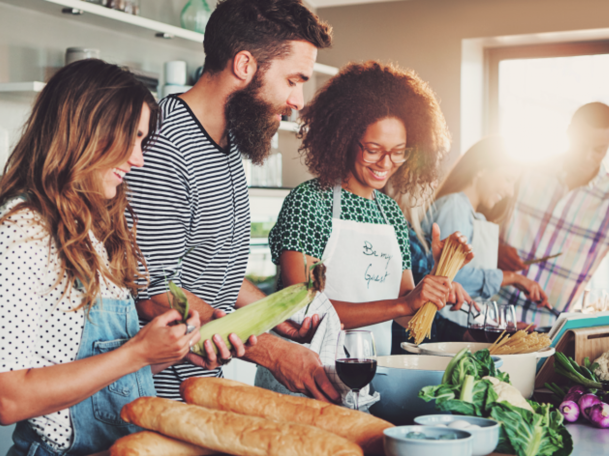 A group of people in a kitchen, preparing food together, with ingredients like bread, corn, and pasta. Sunlight streams through a window in the background.
