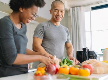 Two people are preparing vegetables together in a bright kitchen while smiling, with various fresh produce on the counter, ending the sentence.