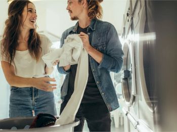 Two people are happily doing laundry together, with one holding a piece of clothing and the other reaching for it, in a bright, modern laundry room.