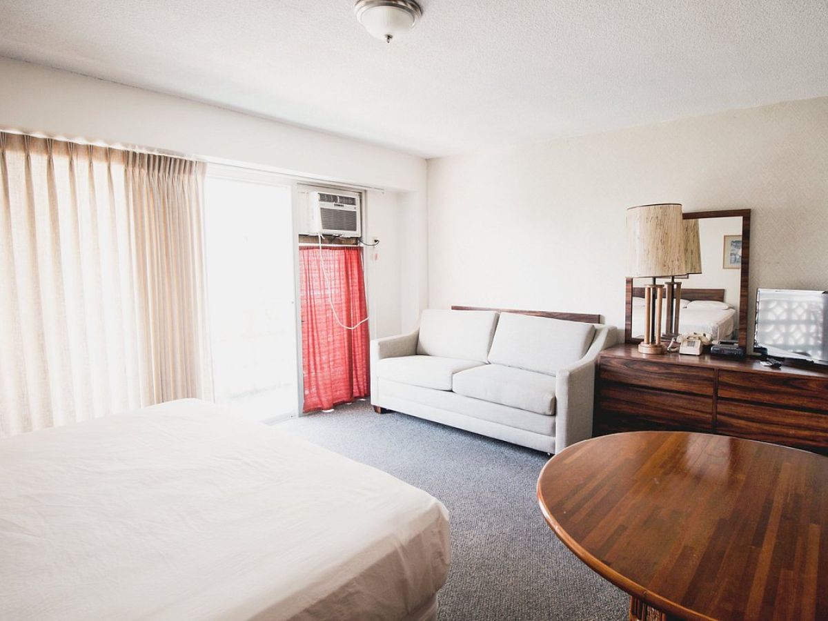 A bedroom with a bed, sofa, round table, TV on a dresser, and window with curtains. There is a lamp and mirror on the dresser.