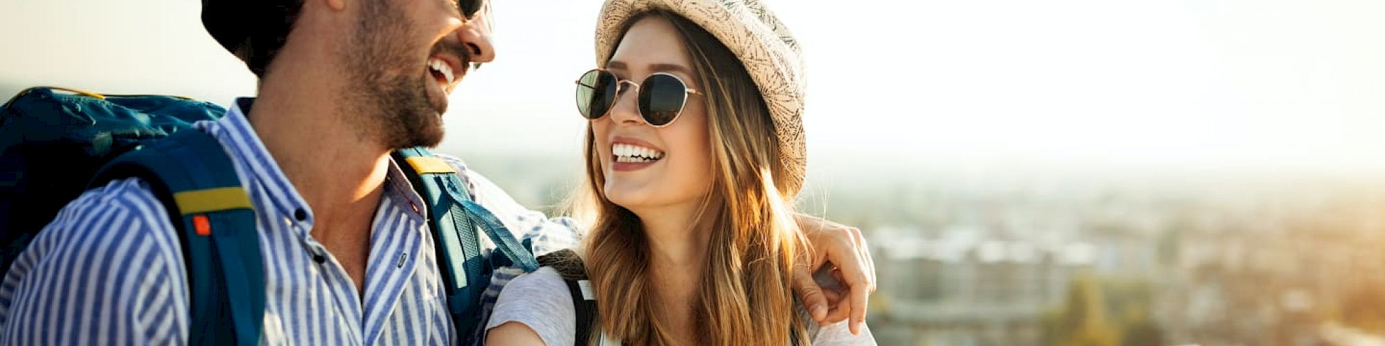 A smiling couple wearing sunglasses and hats, with backpacks, enjoying an outdoor setting while holding a map, suggesting they are traveling.