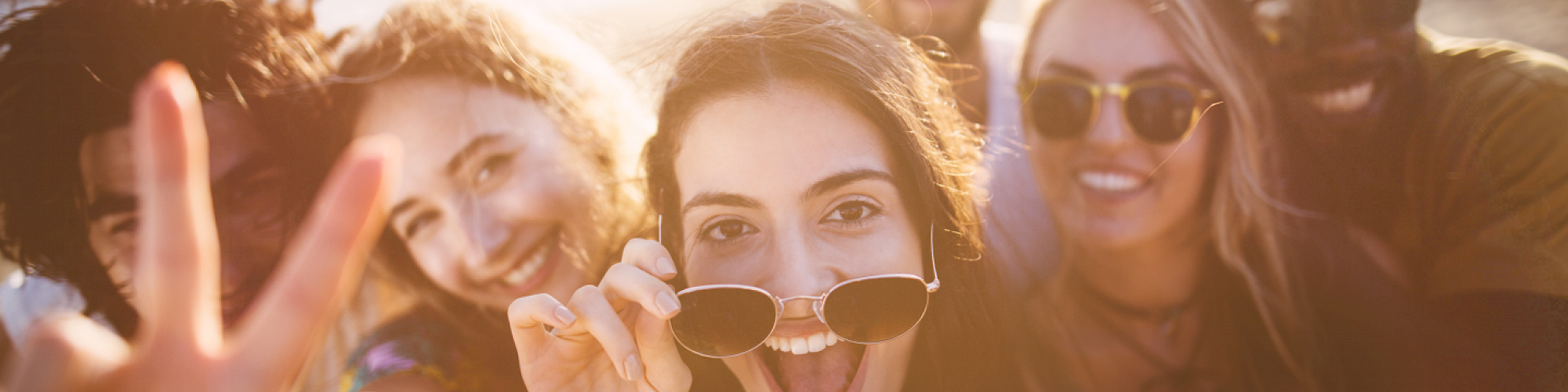 A group of six friends is taking a cheerful selfie outdoors, with some wearing sunglasses and smiling brightly in the sunlight.