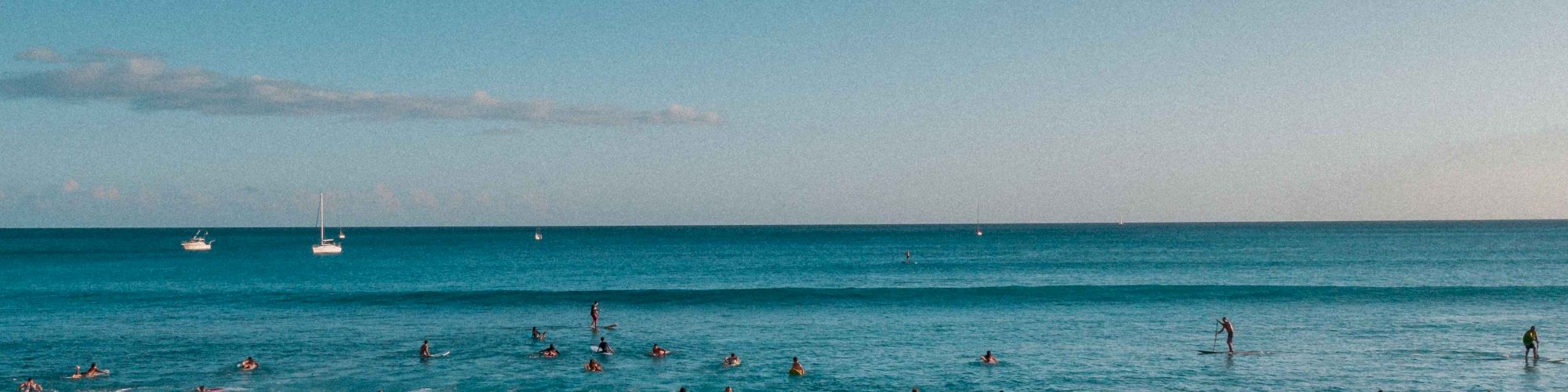 The image shows a group of surfers riding waves in the ocean under a clear blue sky, with sailboats visible in the distance.