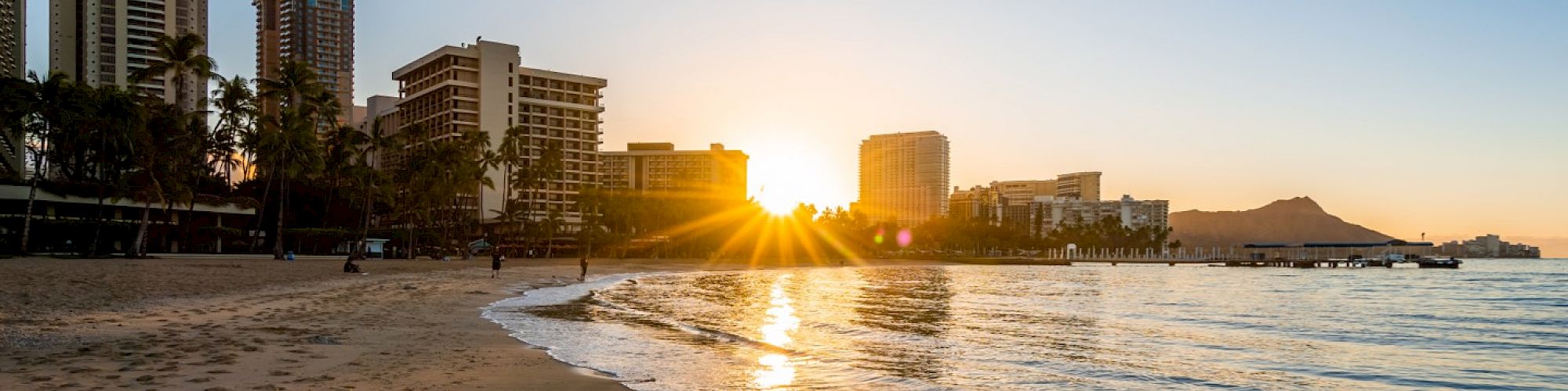 A serene beach at sunrise with the sun peeking over modern buildings and distant mountains, reflecting on the calm water's edge.