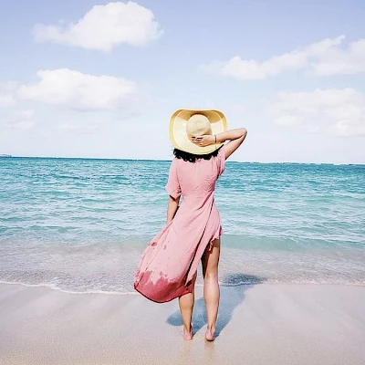 A person in a pink dress and yellow hat stands on a sandy beach facing the ocean with a blue sky and clouds in the background.
