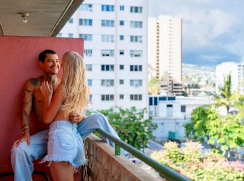 A couple is embracing on a balcony, surrounded by urban buildings and greenery, under a partly cloudy sky.