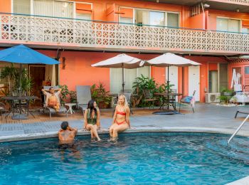 A poolside scene with people relaxing: two sitting on the pool edge, one in the water, and another reading under umbrellas, near a peach building.