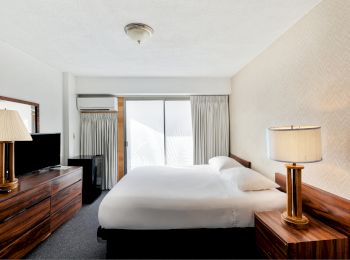 A hotel room with a double bed, wooden furniture, a flat-screen TV, lamps, and a sliding door leading to a balcony.