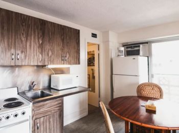 This image shows a small kitchen with a stove, sink, microwave, fridge, and a round dining table with chairs. It has wood cabinets and bright light.