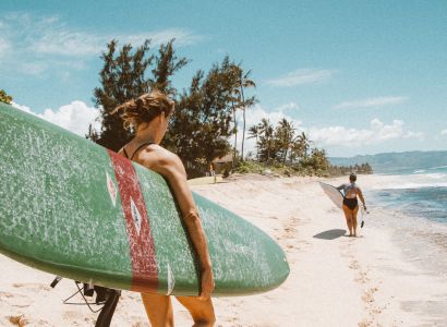 Two people are walking on a sandy beach carrying surfboards. One person leads while the other follows. The sky is clear with a few clouds.