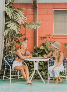 Two people sit at a table outside near surfboards and plants, against a red wall with windows and a door.