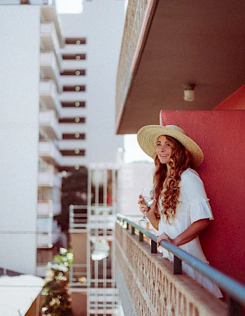 A woman wearing a hat is smiling and leaning on a balcony railing, with a building in the background.