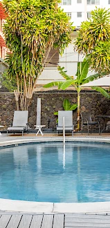 The image shows a circular swimming pool surrounded by lounge chairs, umbrellas, and lush greenery, within a hotel courtyard setting.