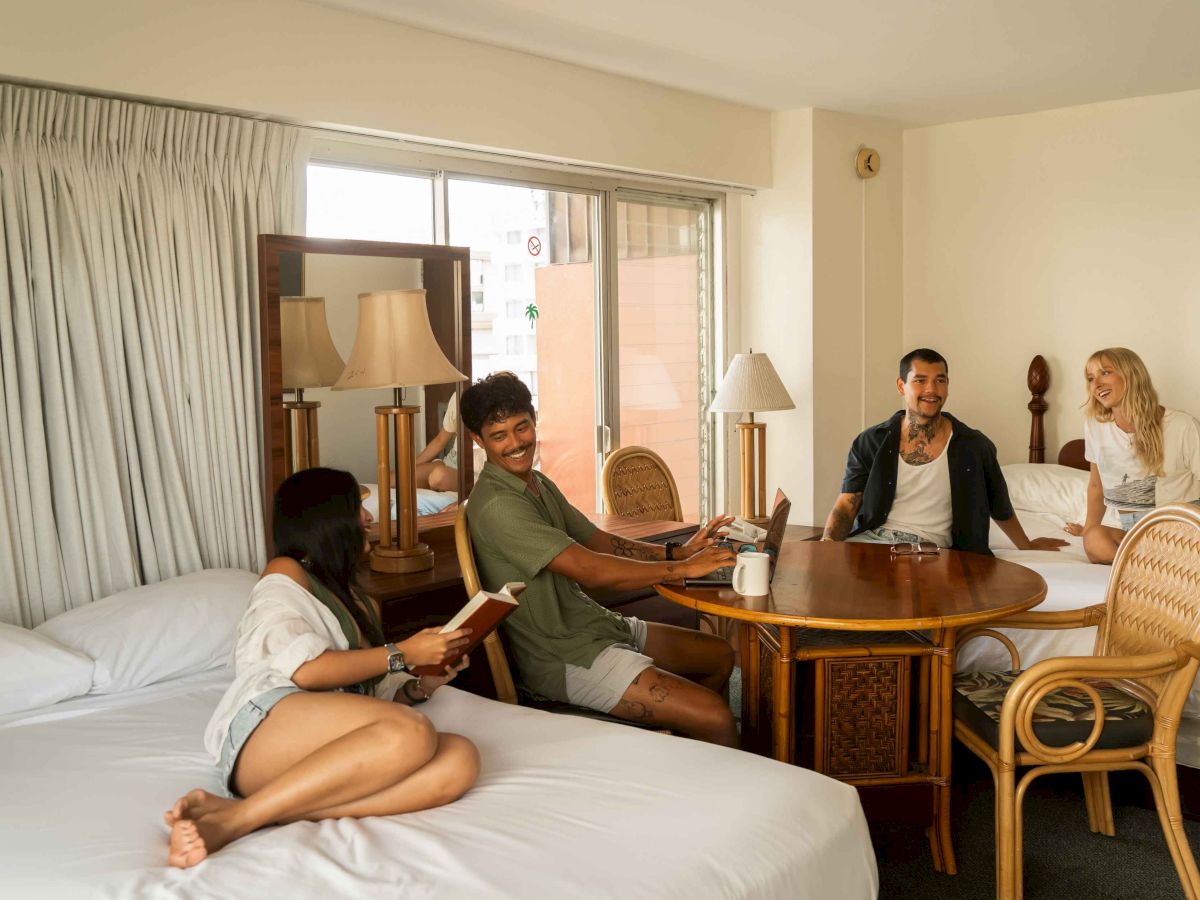 Four people are in a hotel room; two are sitting at a table, and two on a bed, one reading. The room looks bright and comfortable.