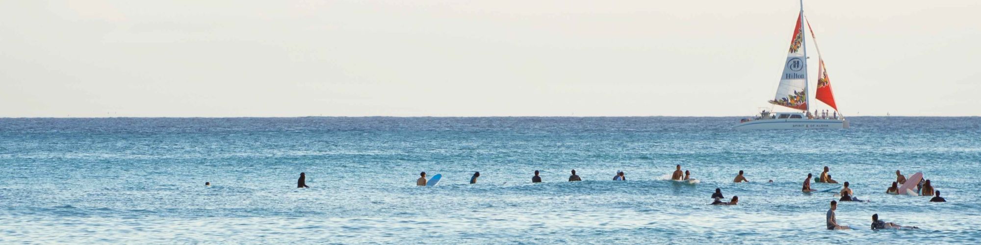People are swimming and surfing in the ocean, with a sailboat in the distance on a clear day.