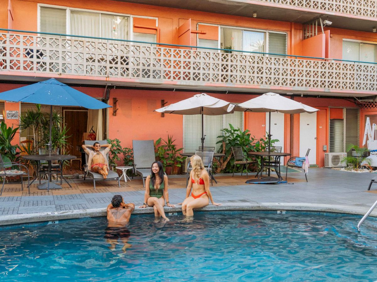 People are relaxing by a pool at a hotel with orange walls and umbrellas, enjoying a sunny day.