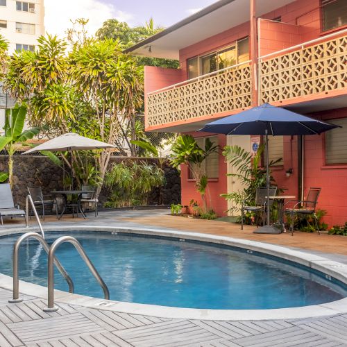 A small, oval swimming pool with lounge chairs and umbrellas is surrounded by a pink building and lush greenery in a sunny area.