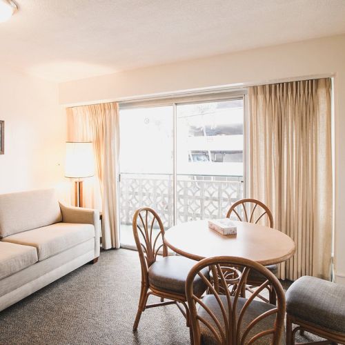 A cozy living area features a beige sofa, a round dining table with four chairs, a standing lamp, and large sliding glass doors leading to a balcony.