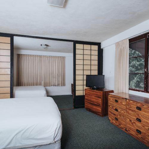 A hotel room with a bed, wooden dresser, television, sliding panel doors, a window with curtains, and an additional bed in the adjoining space.