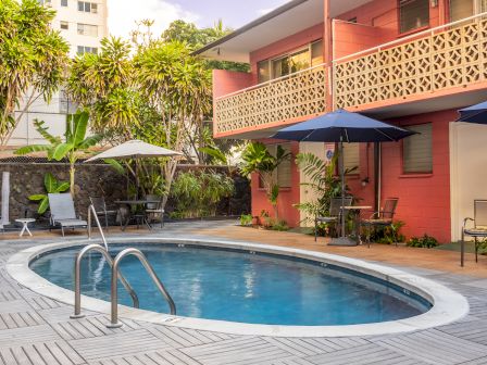 A cozy courtyard features a small oval pool, surrounded by lounge chairs, umbrellas, and a two-story building with a retro aesthetic.