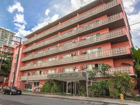 The image shows a multi-story pink building with balconies, likely an apartment or hotel, and a street view with trees and a car parked.