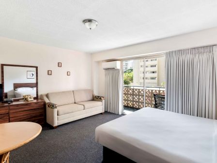 The image shows a hotel room with a bed, sofa, dresser, and balcony, featuring a neutral color palette and modern furnishings.