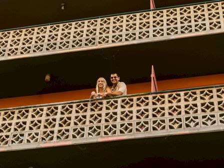 Two people are standing on a balcony with patterned railing, smiling and looking outwards.