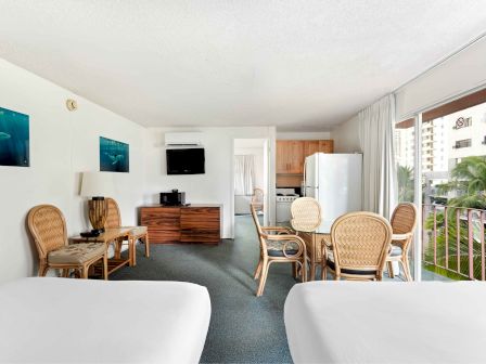 The image shows a hotel room with two beds, a sitting area, a TV, a kitchenette, and a balcony view.