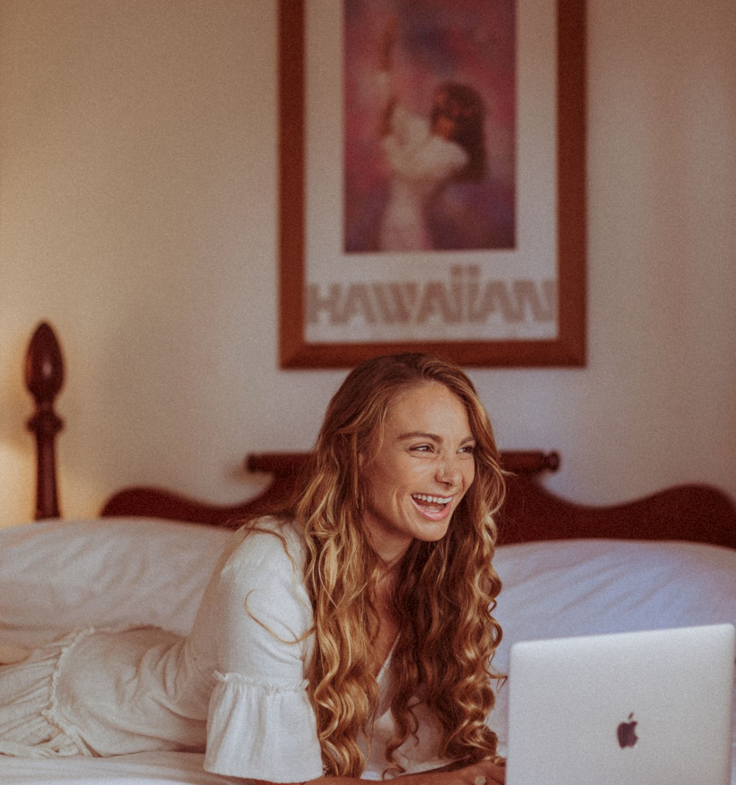 A person with long hair is lying on a bed, smiling at a laptop. A Hawaiian-themed poster is on the wall above the bed.