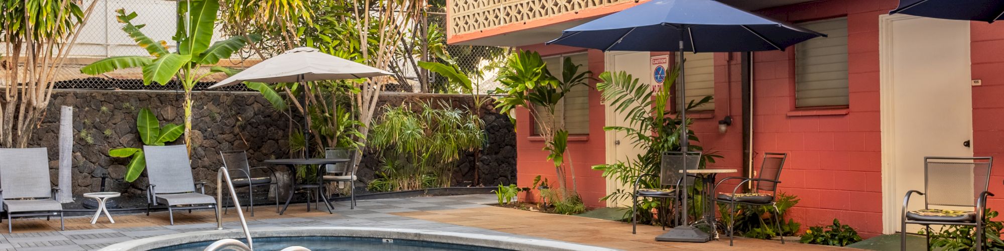 A small, oval pool surrounded by lounge chairs, umbrellas, and tropical plants, next to a building with a patterned railing and pink walls.