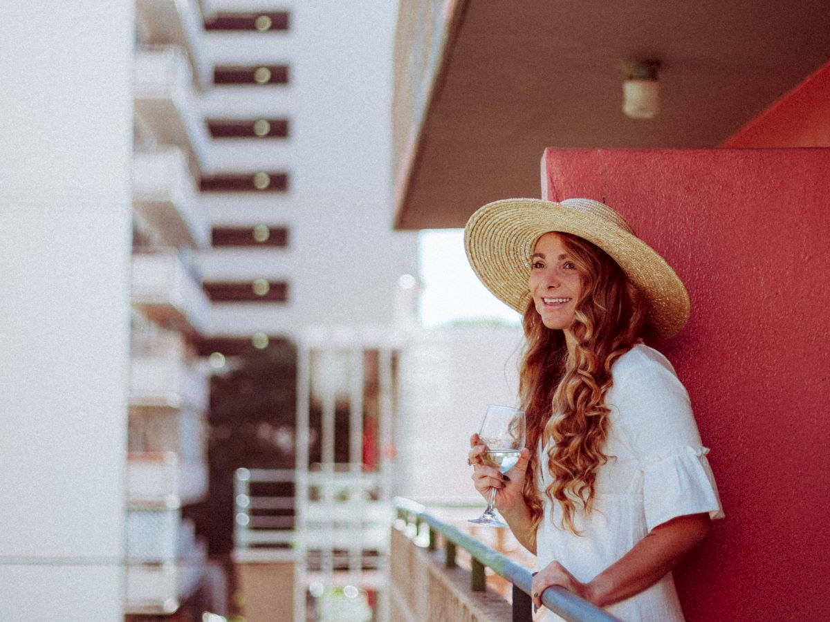 A woman in a straw hat and white dress stands on a balcony of a building, smiling while holding a drink.