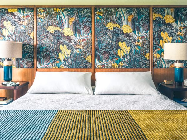 A tidy bedroom with a large bed, vibrant patterned headboard, two bedside tables with lamps, and a colorful knitted blanket on the bed.