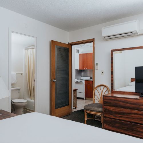 The image shows a tidy hotel room with a bed, dresser, TV, lamp, bathroom, and kitchenette, creating a compact and functional living space.