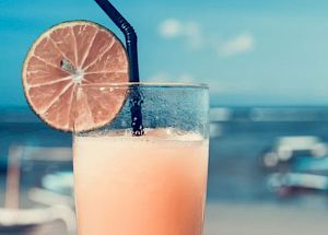 A refreshing drink in a glass with a black straw and a lime slice on the rim, held up with a beach and ocean in the background.